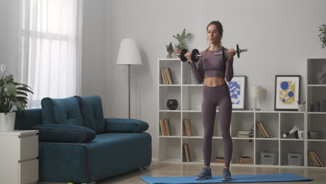 slender athletic woman dressed sportswear is lifting dumbbells training in living room at daytime full-length portrait of sportswoman in modern interior