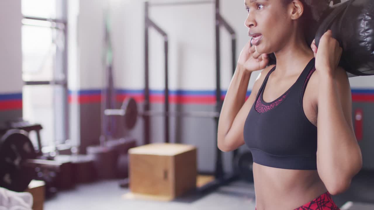 video de una mujer afroamericana decidida haciendo sentadillas sosteniendo un peso de bolsa de arena en un gimnasio.