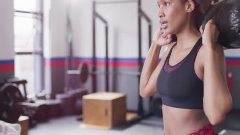 Video-of-determined-african-american-woman-doing-squats-holding-sandbag-weight-at-a-gym