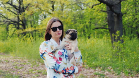 joven mujer elegante con gafas de sol caminando en el parque con un perro de raza pug 2