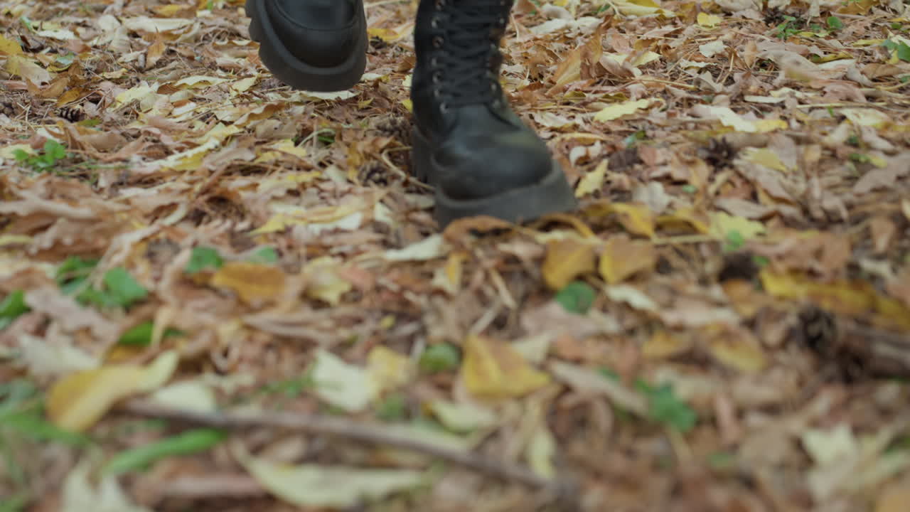 Sepatu bot kulit hitam berjalan melewati dedaunan, close up sepatu bot bertali menginjak-injak dedaunan yang berguguran di lantai hutan, tekstur kasar kulit dan tanah, cahaya musim gugur yang melankolis, langkah pemberontak soliter