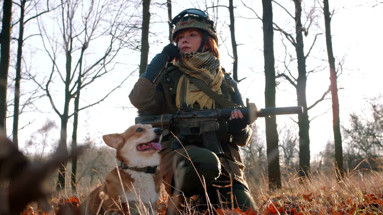 Jeune et jolie femme rousse en uniforme militaire, armée d'un fusil, jouant avec un chien