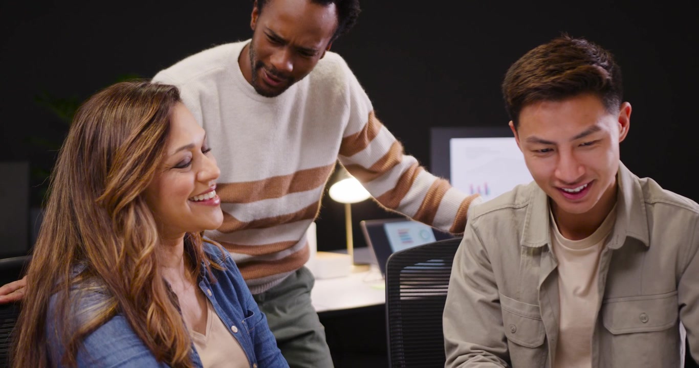 Diverse coworkers at desk, standing man leaning in, leading discussion reviewing monitor charts
