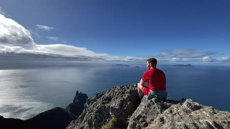 male sits on mountain top, looks out at ocean and blue sky clouds, new zealand