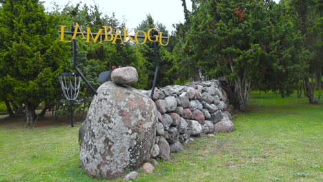 a stone lamb or sheep sculpture in saaremaa near a traditional stone wall holding up a sign that says lambakogu, a popular tourist attraction site in estonia. juniper trees in the background at day.