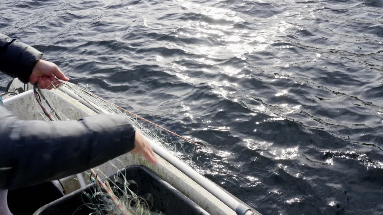 Man retrieves an empty crab net from the sea into a boat, with sunlight glimmering on the calm water, handheld