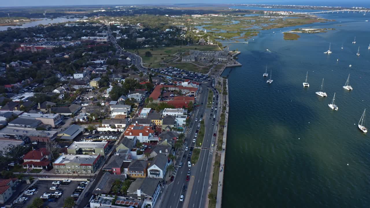 Kuststadslandskap som visar bostadsområden längs en havsmur och väg, med segelbåtar förankrade i lugna vatten och träskmarker som sträcker sig in i fjärran under klar himmel