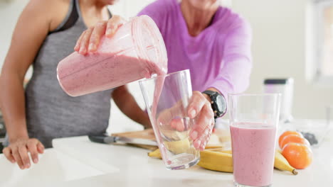 two happy diverse senior women pouring cocktail into glasses in kitchen, slow motion