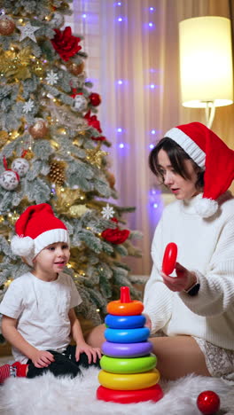 lovely mom and son in red santa caps sit near the christmas tree. happy mother and baby play with pyramid. vertical video.