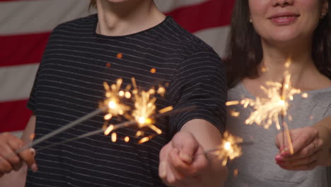 Studioporträt-Einer-Multikulturellen-Gruppe-Von-Freunden,-Die-Vor-Der-Amerikanischen-Flagge-Ein-Wunderkerzenfeuerwerk-Halten-Und-Den-Unabhängigkeitstag-Am-4.-Juli-Feiern-1