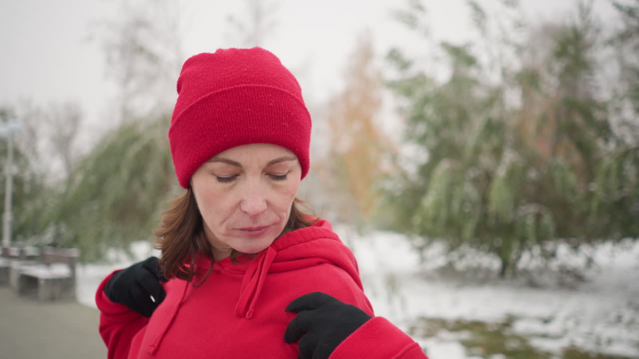 dama con capucha roja con las manos colocadas sobre los hombros gira el brazo y gira lentamente la cabeza hacia la izquierda en un parque cubierto de nieve, el fondo presenta árboles helados, bancos y una atmósfera de invierno serena