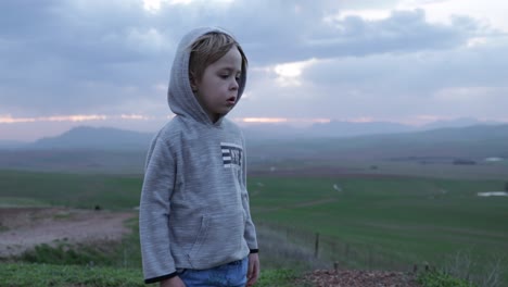 Un-Niño-Solitario-De-4-Años-Con-Una-Mirada-Preocupada-Durante-La-Puesta-De-Sol-Y-Las-Montañas-En-El-Fondo