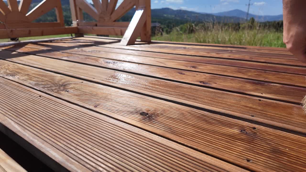 Working man oils varnishing impregnating wooden terrace boards. Decorative railing, serene sky, clouds, and mountain horizon evoke home improvement and outdoor maintenance themes - DIY