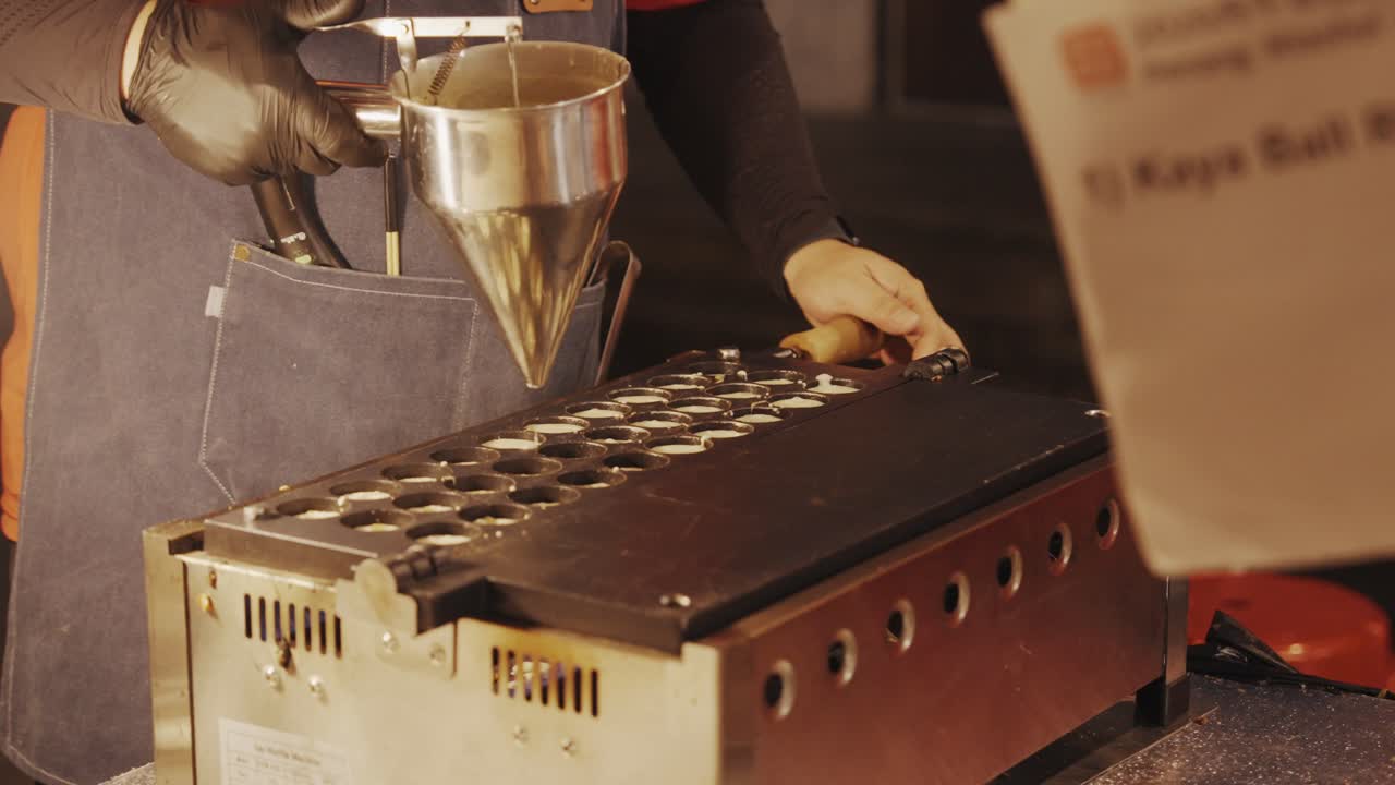 Un primo piano di un venditore di cibo da strada che usa un imbuto da pasticceria per versare la pastella in una cialdiera per waffle a uovo al mercato notturno.