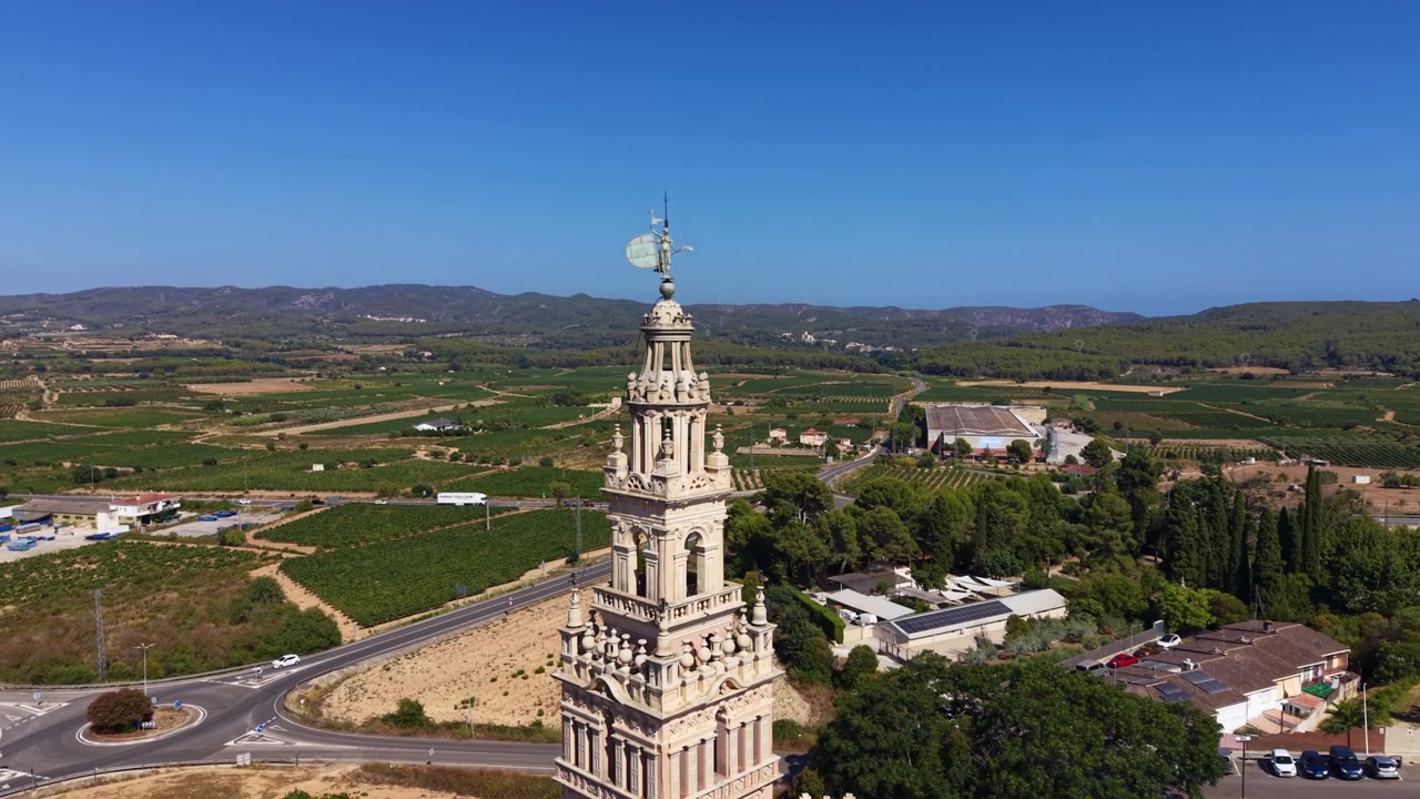 Campanile in pietra ornato di una chiesa si erge sopra un paesaggio rurale spagnolo con vigneti verdi, colline ondulate, una strada circolare e cielo azzurro limpido