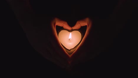 romantic lit heart shaped white candle on black background being held by hands