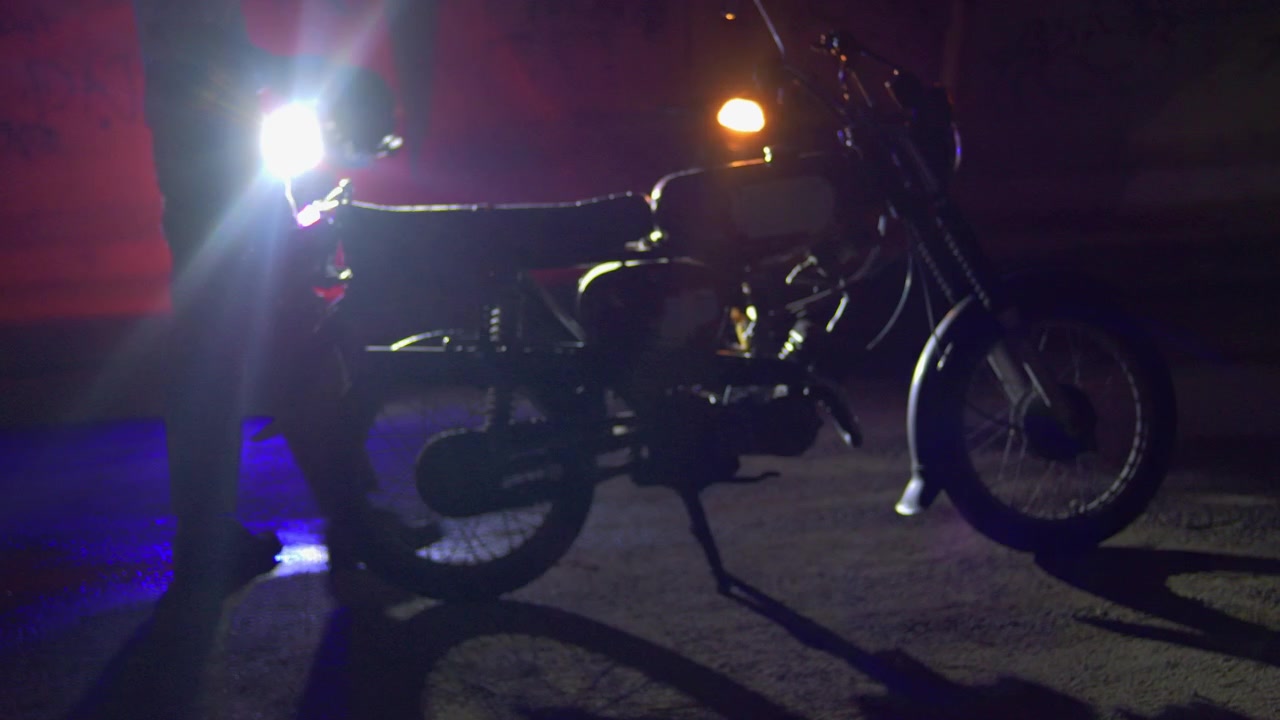 Un uomo cammina verso una motocicletta di notte portando un casco in mano, illuminato da forti fari e luce blu a bordo strada in una scena di viaggio cinematografica e scura.