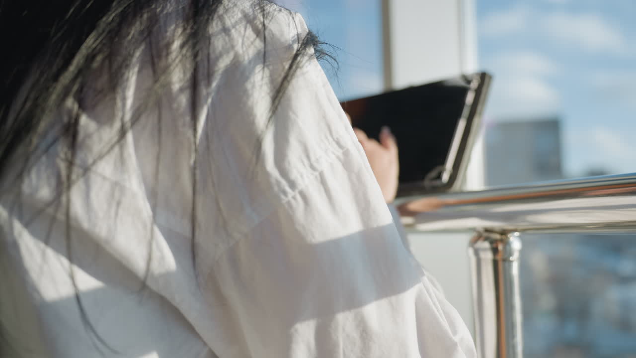 Back view of woman with long straight dark hair standing by glass railing and using tablet with soft urban background and morning sunlight casting gentle reflections on glass and hair strands