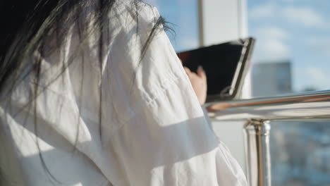 back view of woman with long straight dark hair standing by glass railing and using tablet with soft urban background and morning sunlight casting gentle reflections on glass and hair strands