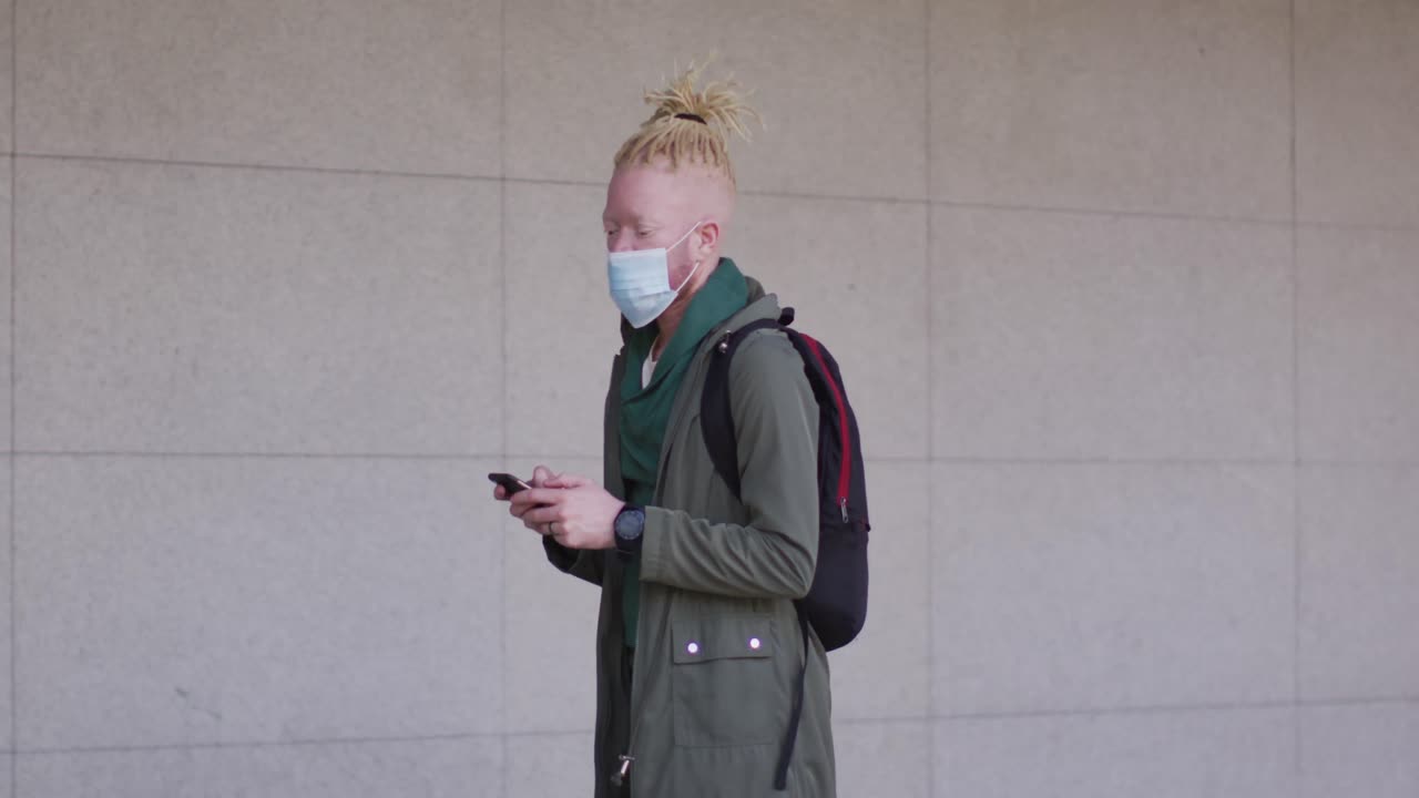 Albino african american man with face mask and dreadlocks walking and using smartphone