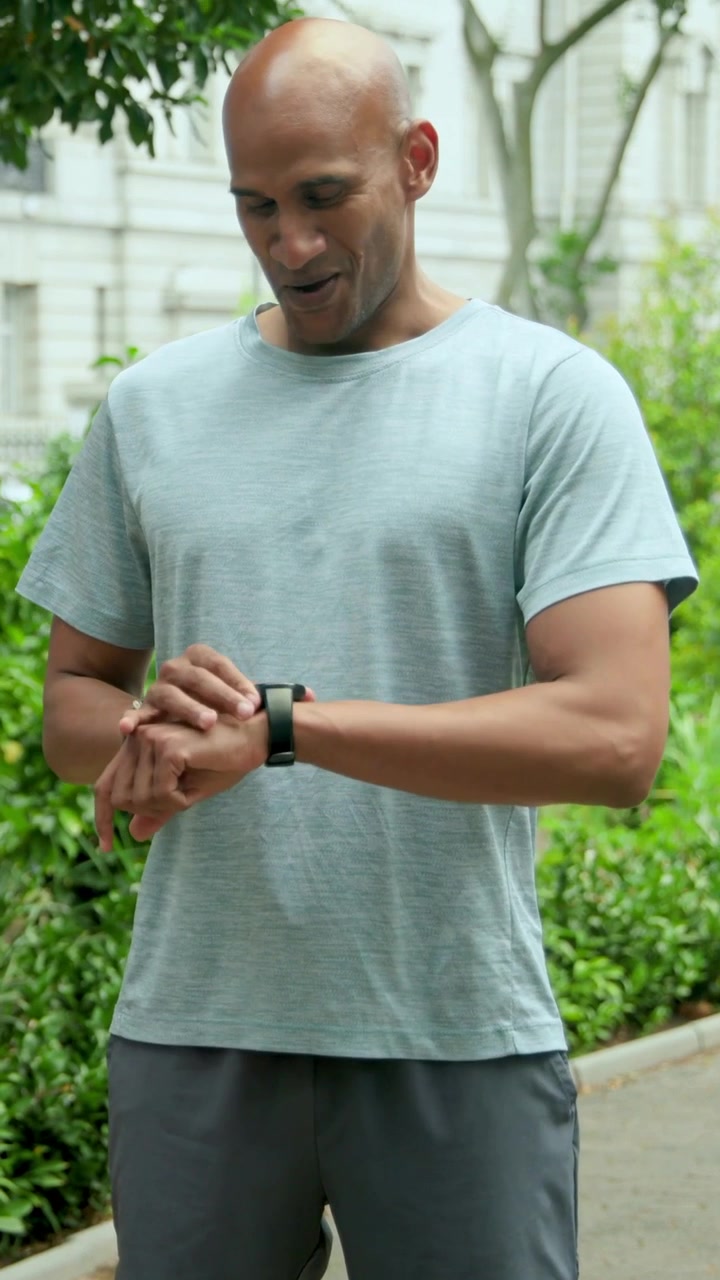 Vertical video: After run panting runner in gray tee dark shorts checking smartwatch on paved path