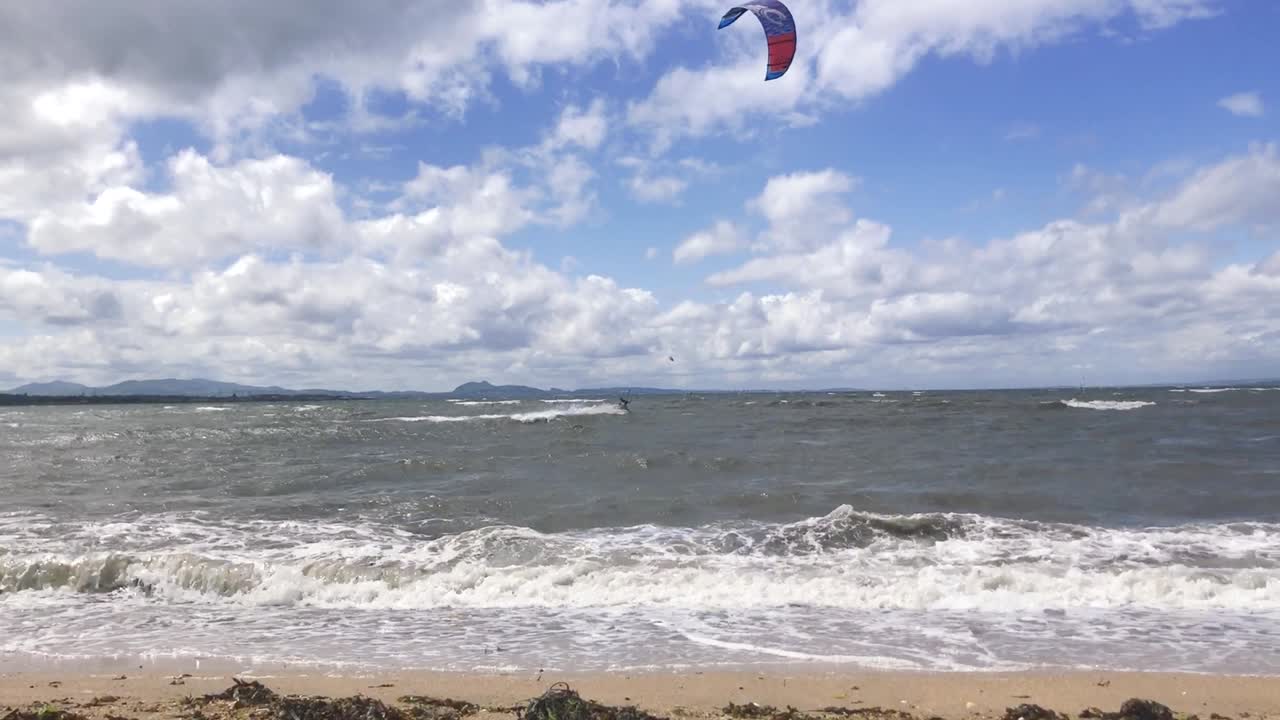 una toma fija de olas rompiendo en una playa de arena con kitesurfistas moviéndose a través de la toma de fondo, en un ventoso día de verano | playa de portobello, edimburgo | filmado en hd a 24 fps cinemáticos