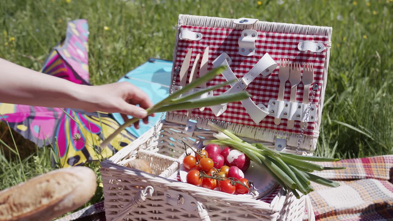A classic wicker picnic basket filled with fresh bread, tomatoes, and vegetables sits on a blanket in a sunny meadow. A person's hand reaches in to take some food