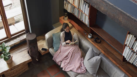 biracial woman lying on sofa under blanket and reading book at home, slow motion