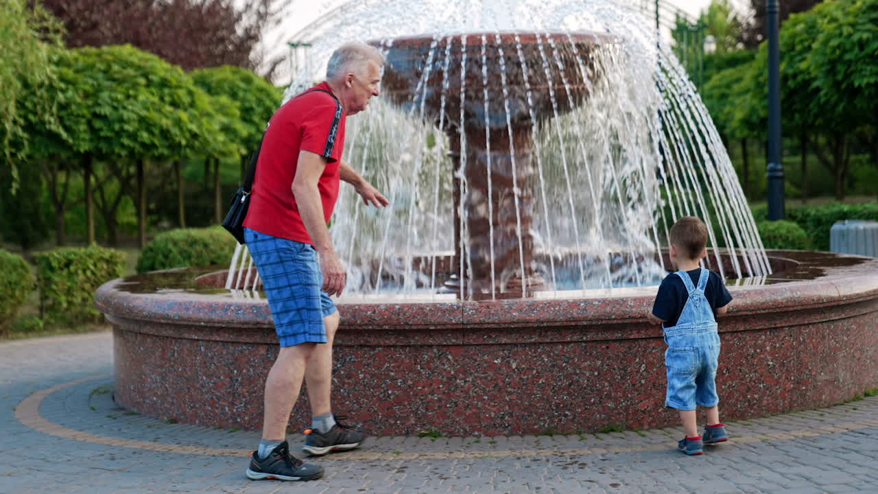 Grandfather with a little boy are in the park. Grandpa touching water in the fountain and grandson follows example.