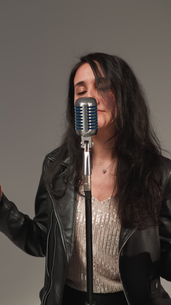A vocalist in a black leather jacket sings with expressive hands and eyes open, standing in front of a vintage microphone against a grey background