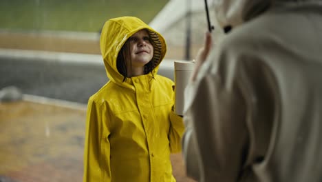 Über-Der-Schulter-Hält-Ein-Glückliches-Junges-Mädchen-In-Einer-Gelben-Jacke-Eine-Flasche-Wasser-Und-Kommuniziert-Mit-Ihrer-Mutter,-Während-Sie-Im-Regen-Im-Park-Spazieren-Geht