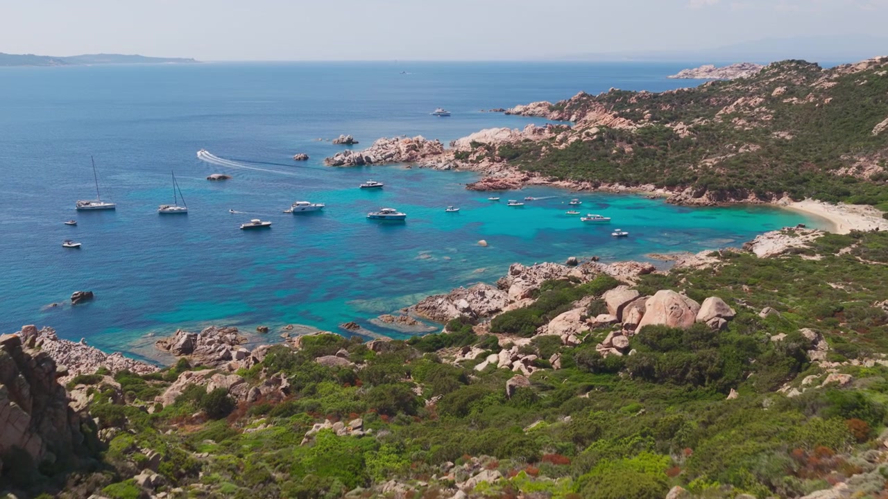 Vue aérienne épique survolant des falaises de granit pour révéler des yachts à voile de luxe dans les eaux bleu turquoise d'un port naturel protégé sur l'île de Spargi, dans l'archipel de la Maddalena en Sardaigne, Italie