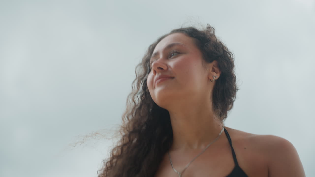 Ritratto ravvicinato di donna multirazziale su una spiaggia isolana, sguardo contemplativo verso il cielo nuvoloso, lunghi capelli ricci mossi dalla brezza, dettaglio della spallina del bikini e della collana, luce naturale soffusa, emozione sottile, viaggiatrice