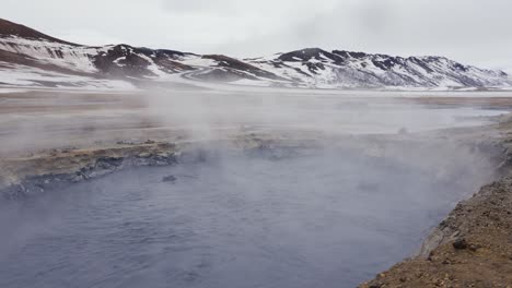 Dark-blue-mud-pool-bubble-and-form-white-vapor-during-bright-day,-Iceland