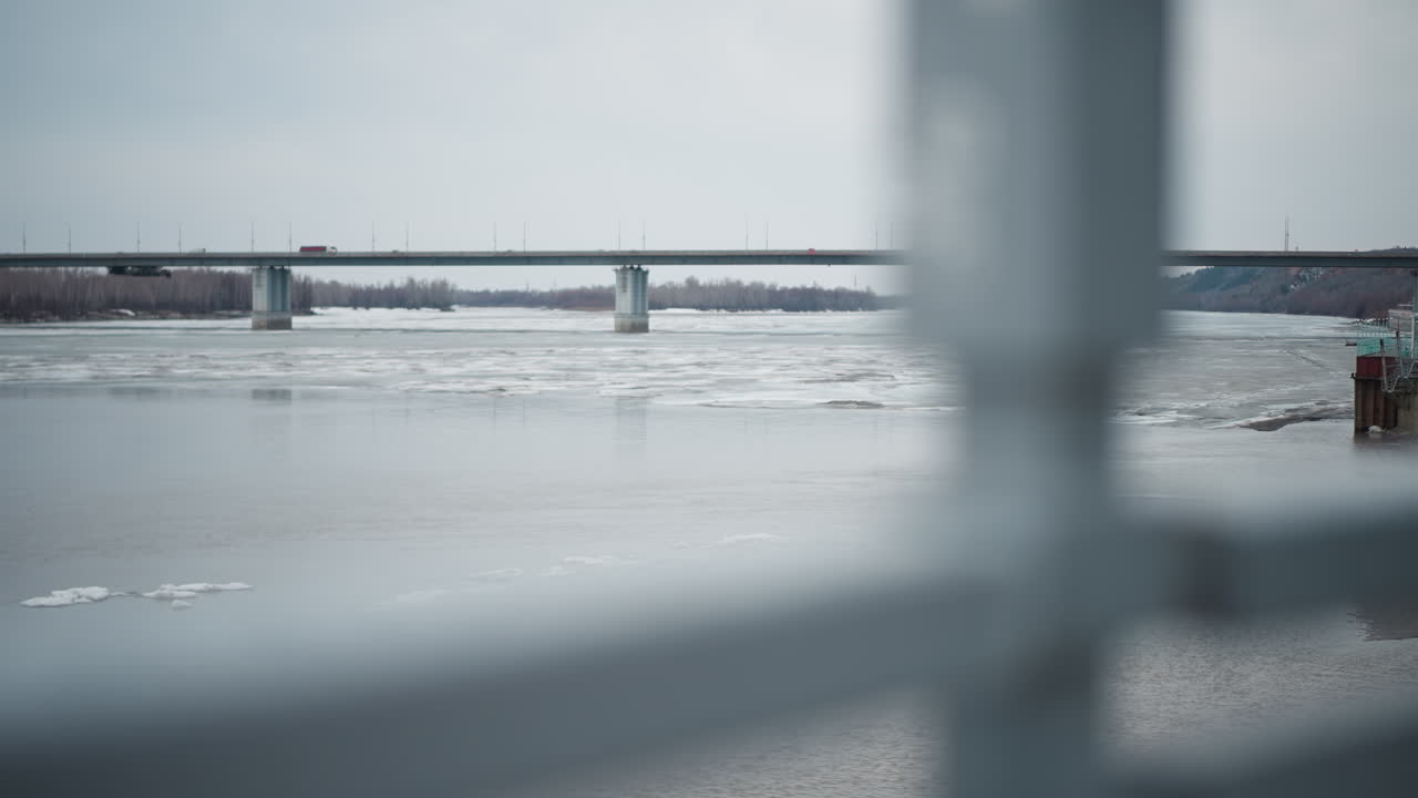 View of frozen river with floating ice chunks under long bridge in overcast weather, framed by blurred metal railing in foreground, distant shoreline with bare trees and hills visible in background