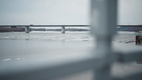 view of frozen river with floating ice chunks under long bridge in overcast weather, framed by blurred metal railing in foreground, distant shoreline with bare trees and hills visible in background