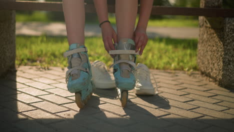 vista de la pierna de la correa de ajuste individual en patines de rodillos de color cian mientras está sentado en un banco con un brazalete negro en la mano derecha, zapatillas blancas descansando detrás, la luz solar cálida proyectando sombras