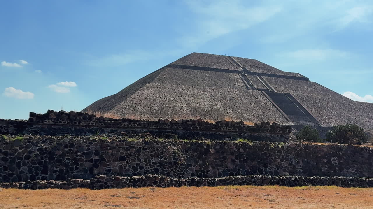 Vista dal basso delle terrazze e dei gradini in pietra della Piramide del Sole di Teotihuacan sotto la luce intensa del sole, ombre drammatiche contro un cielo azzurro limpido.