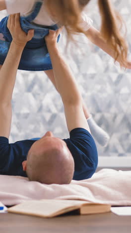 young bald dad plays with his little daughter lying on floor, lifted her in his arms and twist and turn at altitude, long blond hair fluttering, smile, near sunlight