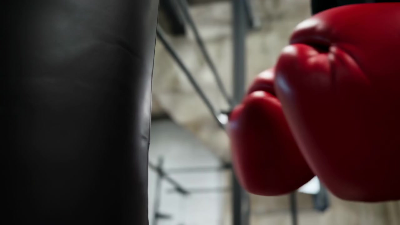 Nahaufnahme zeigt rote Boxhandschuhe, die auf einen dunklen Boxsack drücken, neben einem angeschnittenen Unterarm in einem verschwommenen Fitnessstudio-Interieur, mit hellen Wänden, hängendem Licht und energiegeladener Trainingsatmosphäre, mit Boxen und Boxhandschuh