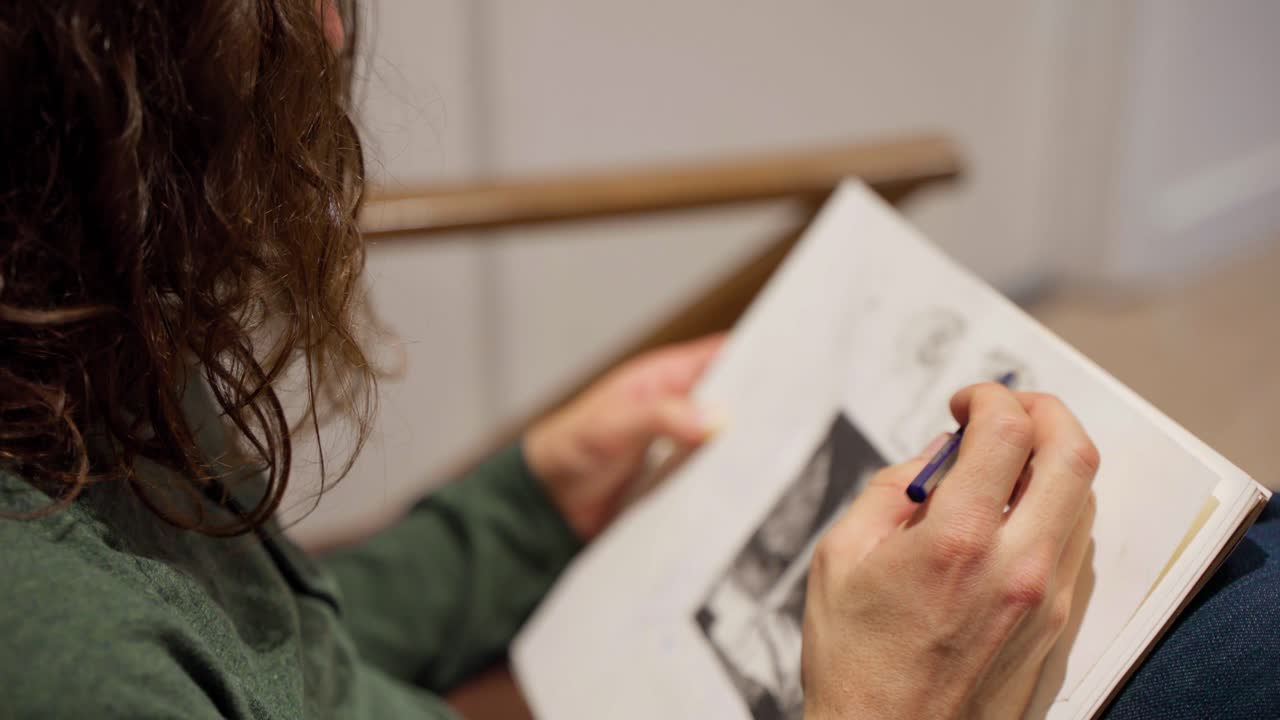 Inquadratura di profilo di un artista con lunghi capelli e maglietta verde che disegna un volto su un blocco da disegno con una matita, sul divano