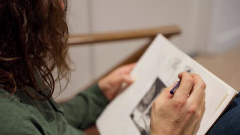 Profile-shot-of-artist-with-long-hair-in-green-shirt-drawing-a-face-on-sketch-pad-with-pencil-on-sofa