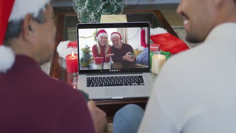 Padre-E-Hijo-Birraciales-Con-Gorros-De-Papá-Noel-Usando-Una-Computadora-Portátil-Para-Una-Videollamada-Navideña-Con-Una-Pareja-En-La-Pantalla