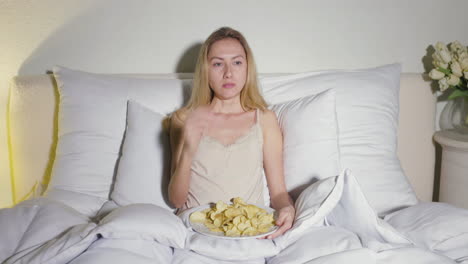 blonde girl sitting in the bed and eating potato chips