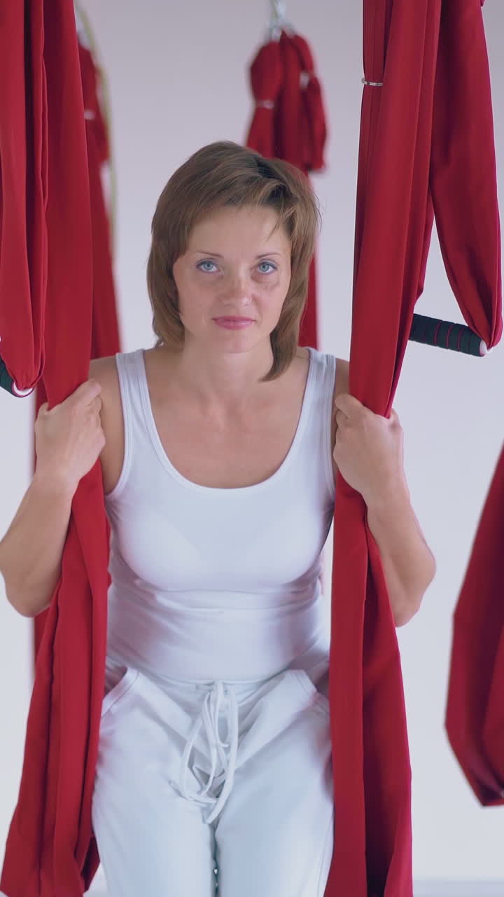 nice blonde woman in white sport suit relaxes after flying yoga workout sitting and swinging on red hammock