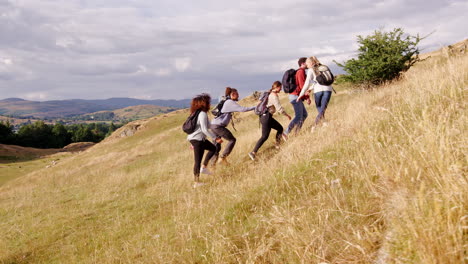 A-multi-ethnic-group-of-five-young-adult-friends-help-each-other-while-hiking-uphill-across-a-field-to-the-summit-of-a-mountain,-tracking-shot