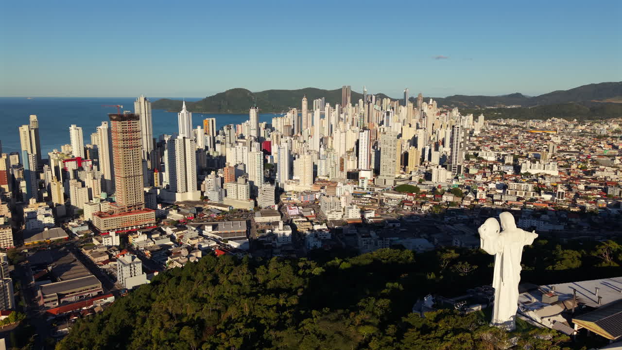 Smooth right-slider drone shot unveils Camboriú’s hilltop Christ statue, framed against morning-lit skyscrapers and ocean background. Ideal for spiritual or destination marketing