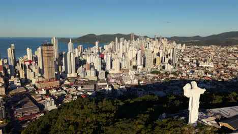 smooth right-slider drone shot unveils camboriú’s hilltop christ statue, framed against morning-lit skyscrapers and ocean background. ideal for spiritual or destination marketing
