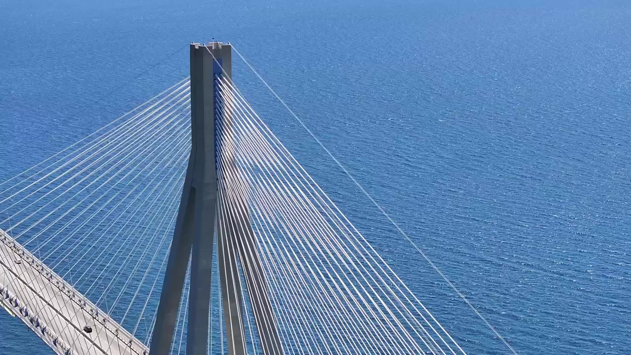 High angle close-up of a massive cable-stayed bridge pylon and intricate stay cables against a serene blue coastal sea.