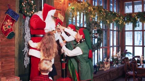 santa claus with firewood in his hands enters the house. joyful children and elves run to santa and hug